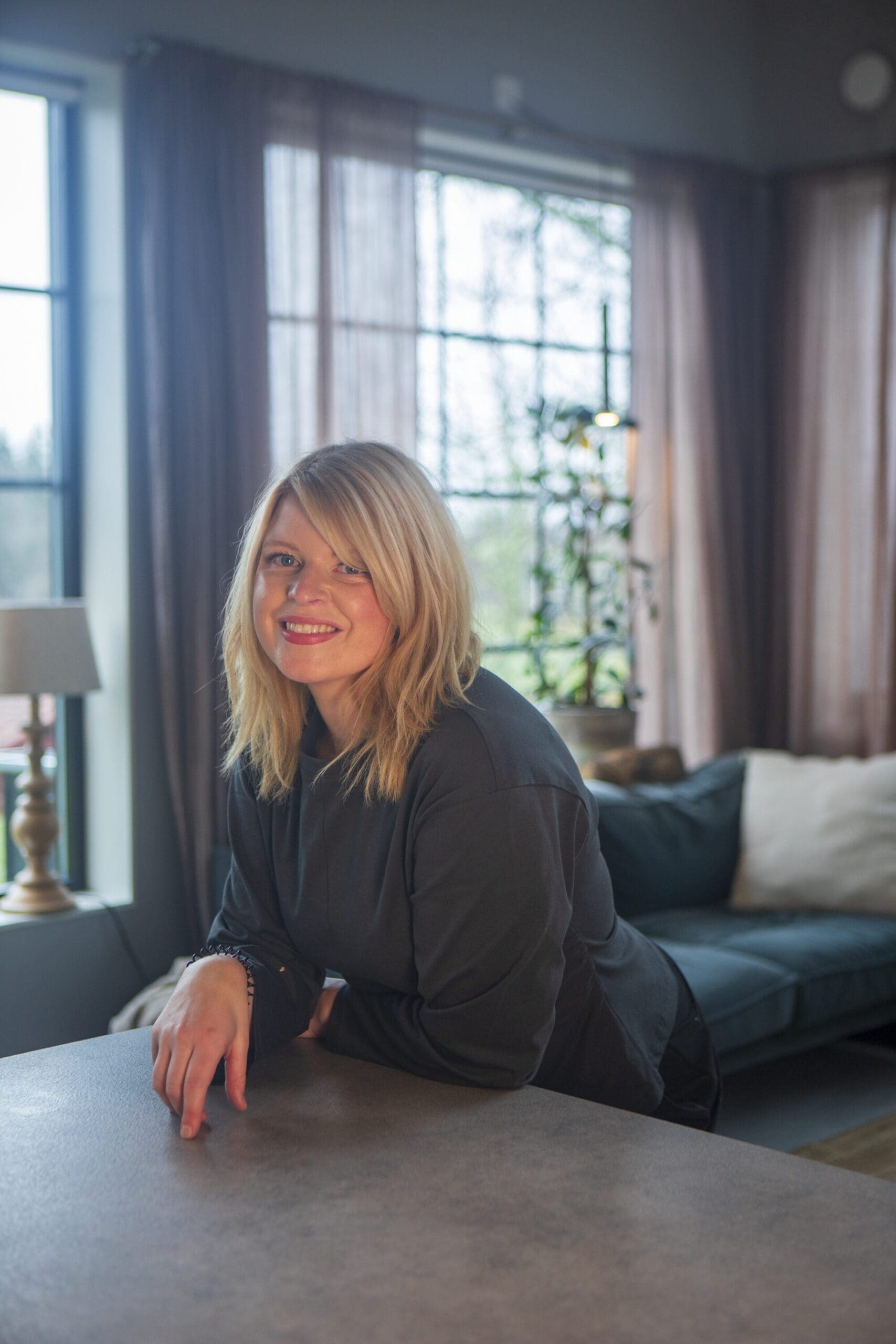 En leende blond kvinna som står vid ett bord. I bakgrunden finns höga fönster.