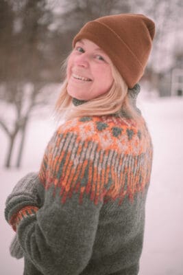 En blond kvinna i en grön stickad tröja och brun mössa som står med ryggen mot kameran och tittar in leende i kameran. Det är snö på marken. Hon har menstrosor på sig.